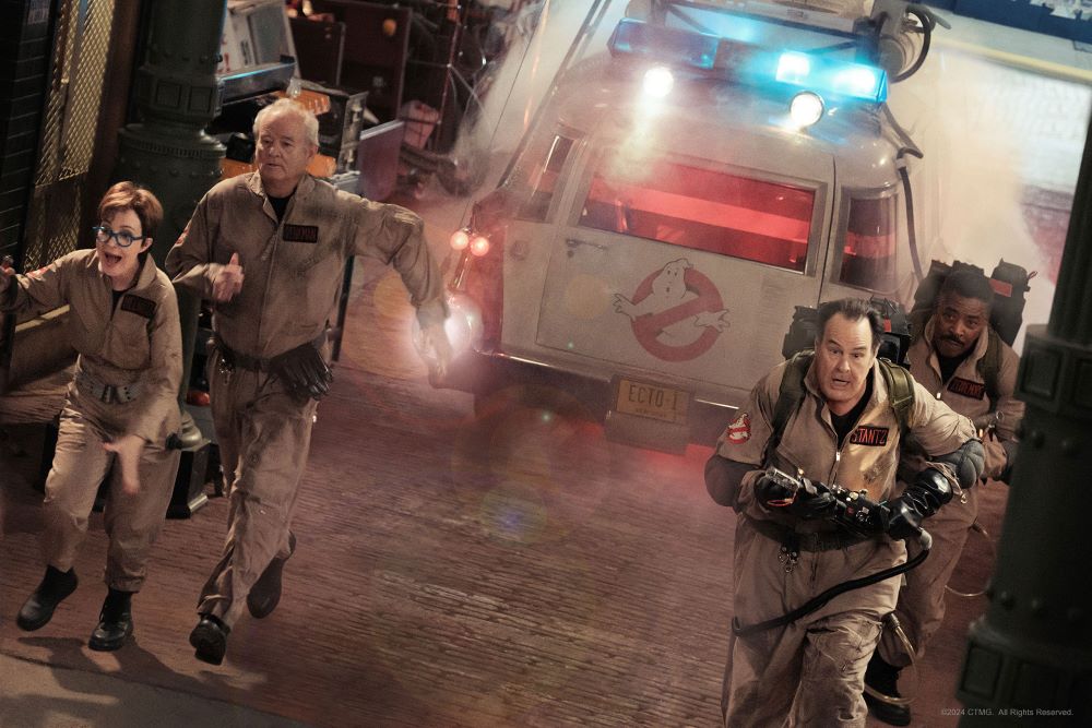 Phoebe and the original Ghostbusters, clad in their uniforms, run through a street while the Ectomobile is parked behind them.
