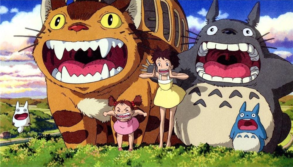 A photo still from the Studio Ghibli film My Neighbor Totoro. Two young girls with brown hair stand beside each other while shouting in a meadow with two large creatures and one small creature surrounding them. 