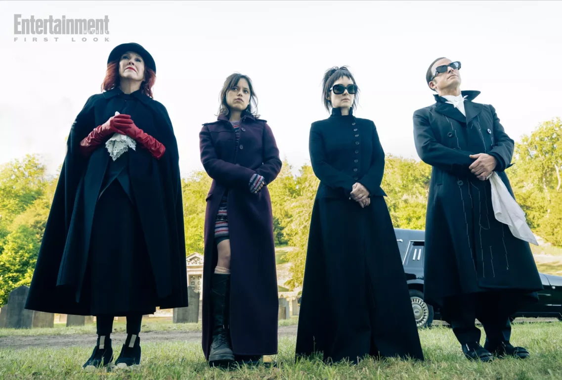 Catherine O'Hara, Jenna Ortega, Winona Ryder and Justin Theroux stand in a line on the grass. All four are dressed in black. There is a hearse behind them.