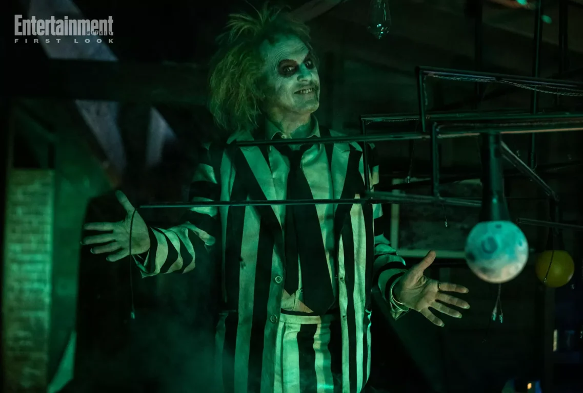 Michael Keaton in character as Beetlejuice. He's dressed in a black and white striped suit and wearing grease paint on his face. Everything is tinted a sickly green.