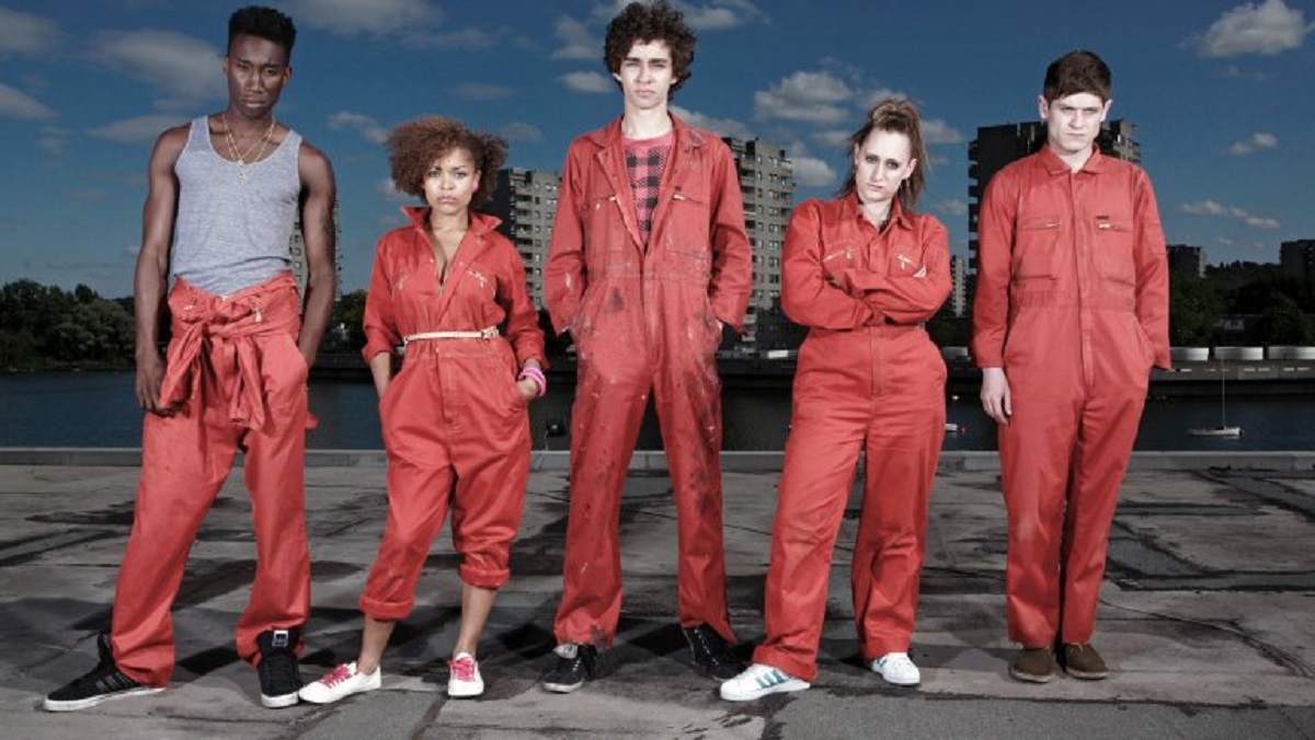 The cast of Misfits stands outside while wearing orange jumpsuits and looking serious. 