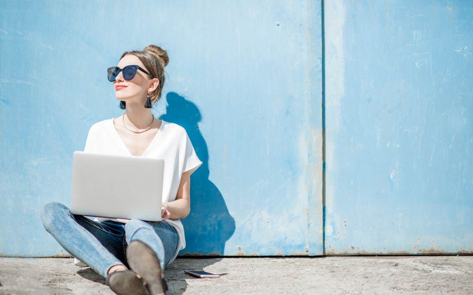 a woman in sunglasses sitting in the sun with a laptop