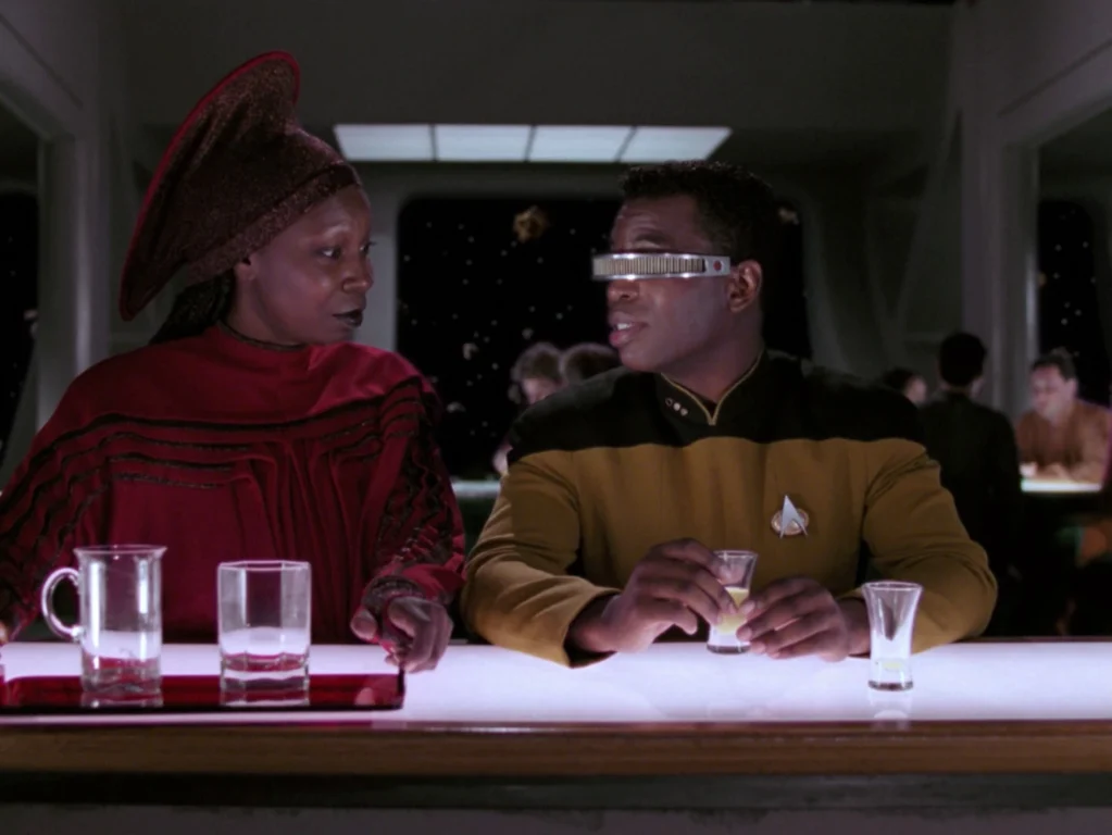 Guinan sets a tray of glasses down on the bar next to Geordi, who has a glass in his hand.