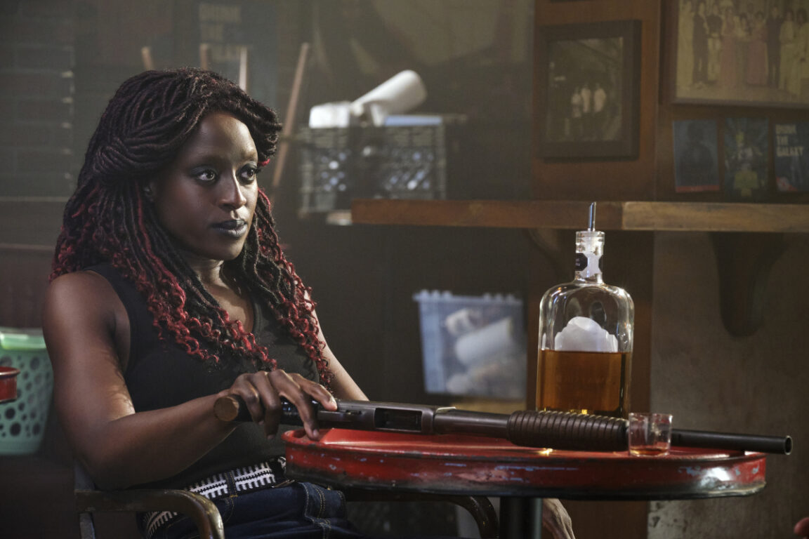 Ito Aghayere as young Guinan, sitting in a restaurant with a weapon on the table in front of her 