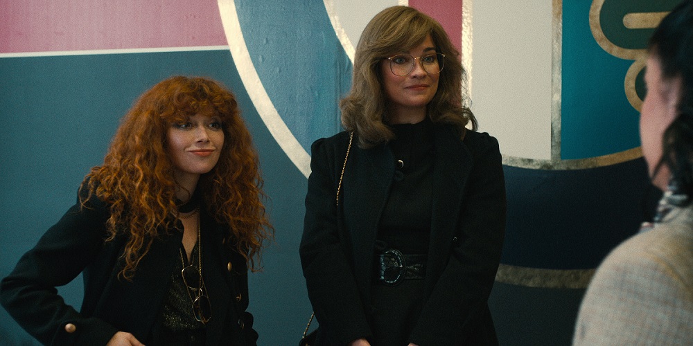 Natasha Lyonne as Nadia and Annie Murphy as young Ruth, standing with a blue, white, and pink wall behind them on Russian Doll Season 2 Episode 2 "Coney Island Baby."