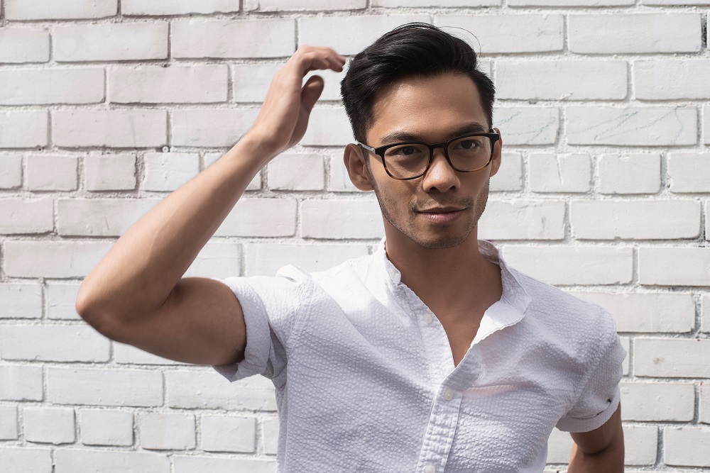Photo of actor Aldrin Bundoc, wearing a white shirt with glasses with a white brick backdrop.