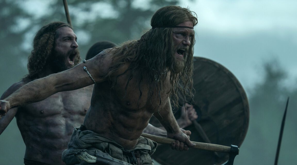 A blonde, long haired viking runs into battle holding a shield. He is played by Alexander SkarsgÃ¥rd in The Northman.