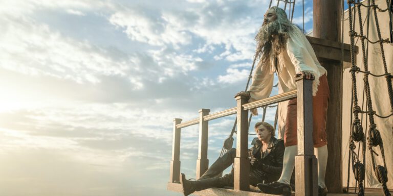 Taika Waititi as Blackbeard and Rhys Darby as Stede Bonnet in Our Flag Means Death. Stede sits and Blackbeard stands against the rail of a ship. They're wearing each other's clothes