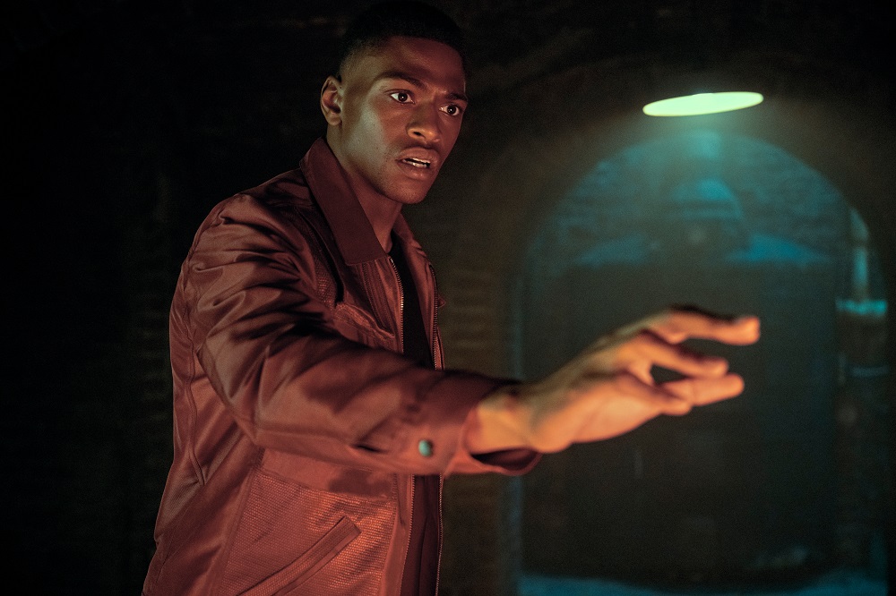 Marcus wearing a red shirt while holding his hand over something in a dark room in The Umbrella Academy Season 3 Episode 1 "Meet the Family."