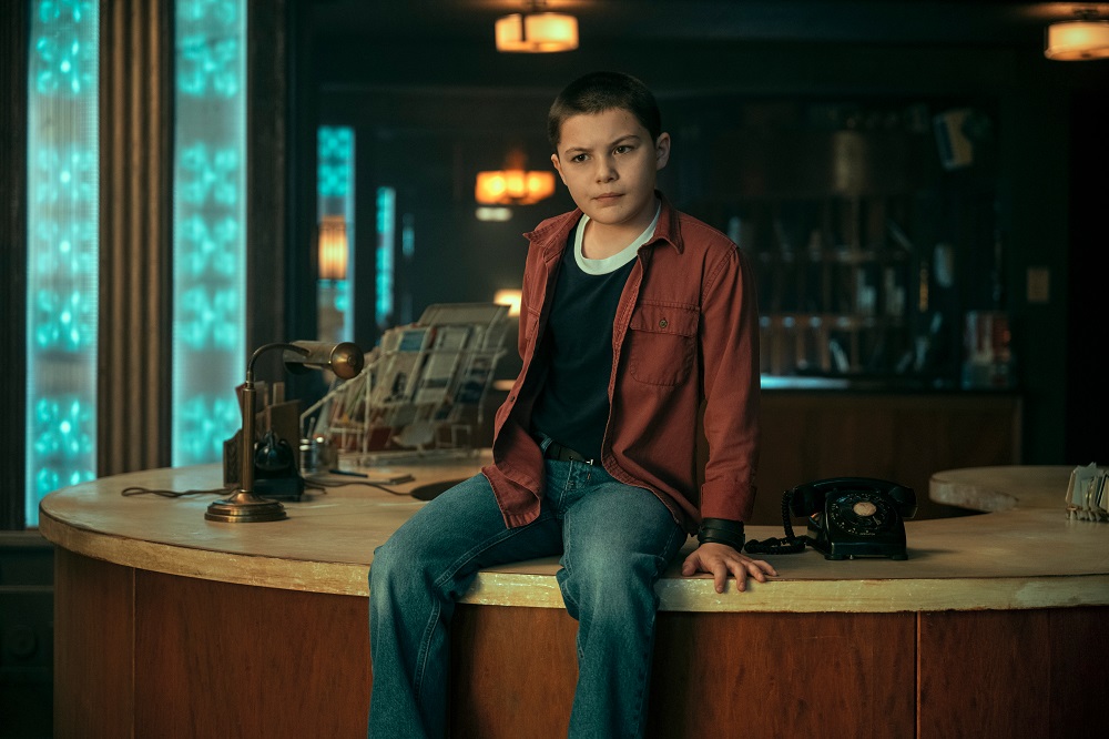 Stan sitting on a receptionist desk while wearing a red shirt in The Umbrella Academy Season 3 Episode 3 "Pocket Full of Lightning."