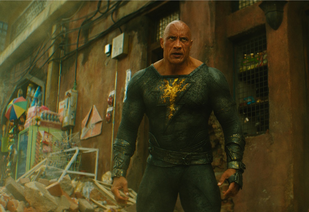 Black Adam stands among debris and rubble in a street while looking angry.
