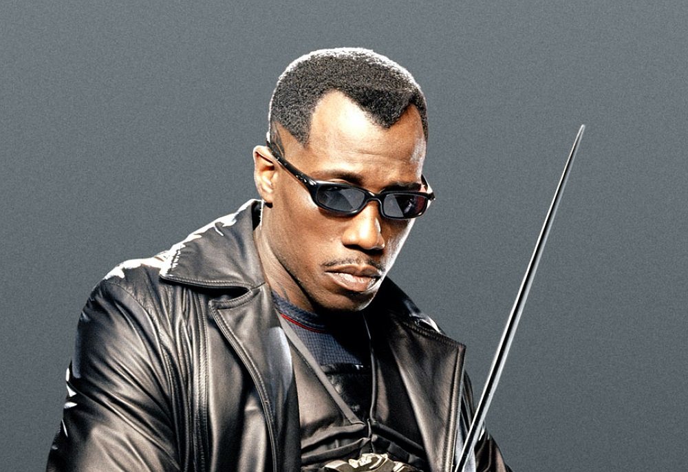 Wesley Snipes as Blade, wearing black sunglasses, a black trench coat, and wielding a sword on a dark gray background.
