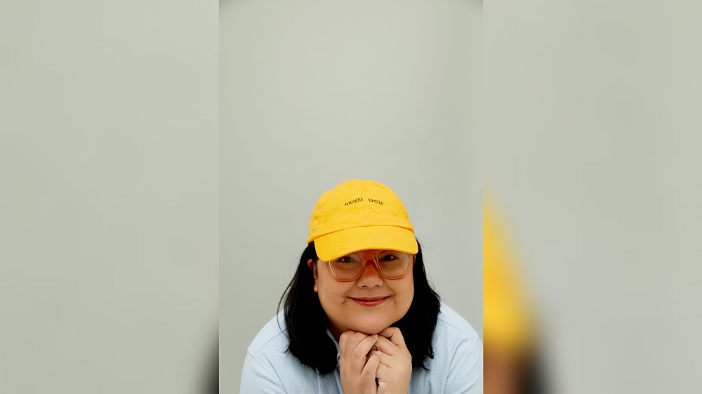 A promo photo for "Moro Girl," a solo comedy show by Alia Ceniza Rasul, featuring Alia wearing a white shirt and yellow hat with an off-white background.