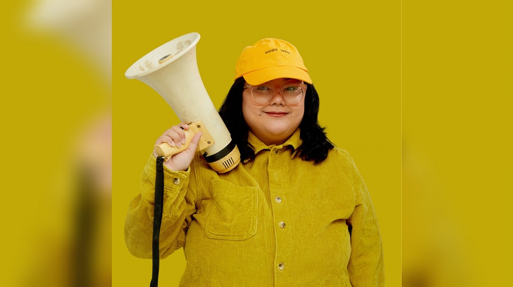 Promo photo for "Moro Girl," a solo comedy show from Alia Ceniza Rasul, with Alia standing while holding a megaphone and wearing yellow with a yellow background.