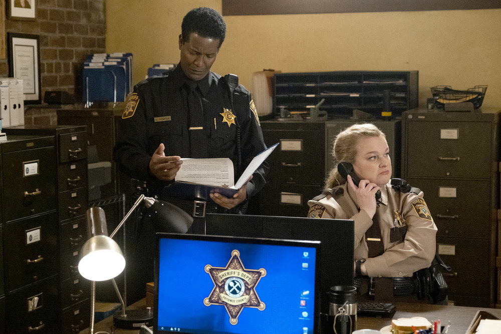 Sheriff Mike Thompson stands over Deputy Liv Baker while he holds a book and she's on the phone on Resident Alien Season 2 Episode 10 "The Ghost of Bobby Smallwood."