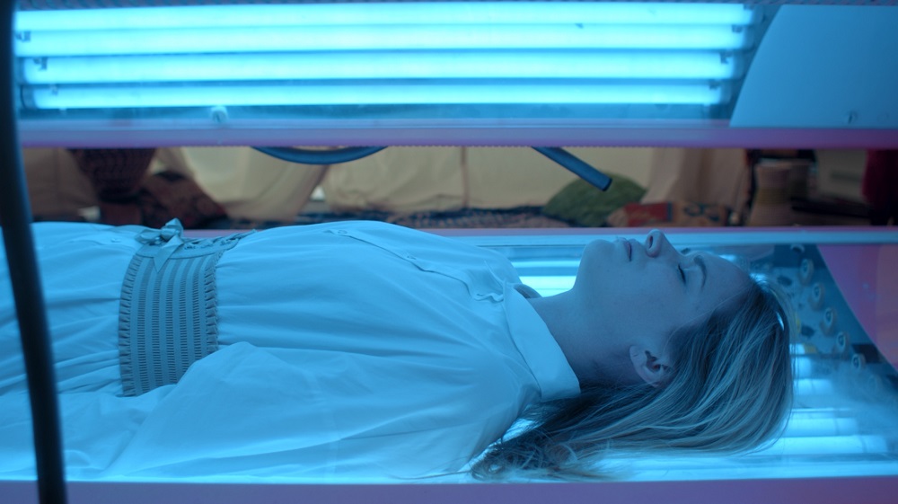 Mack lies in a tanning bed while wearing a white blouse in Mack & Rita.