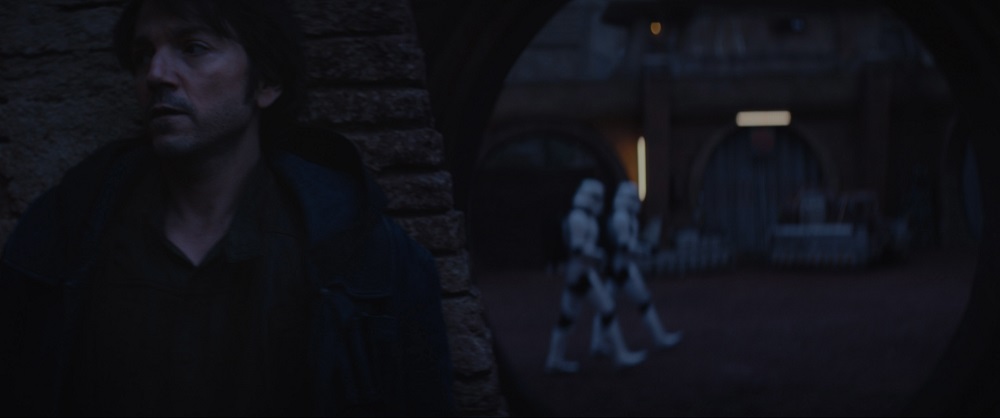 Cassian Andor stands with his back against the wall in hiding while Stormtroopers walk past him.