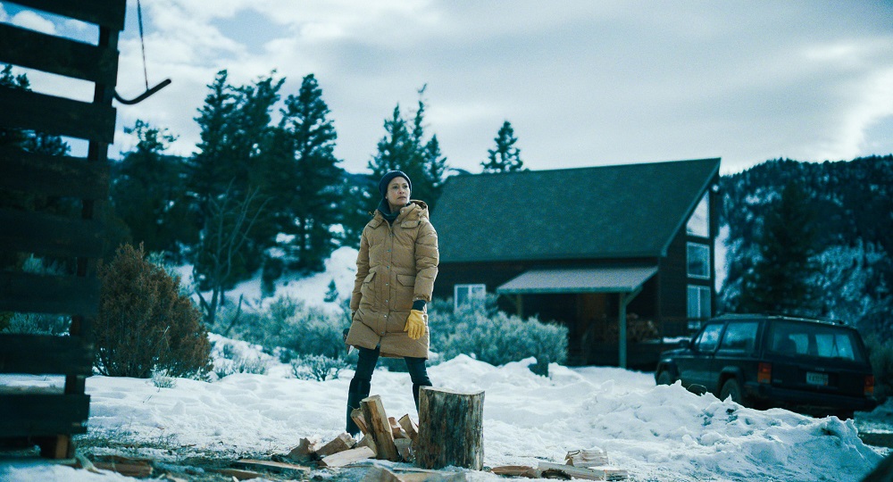 Sandra stands in front of a split log in the snow with her house behind her in God's Country.