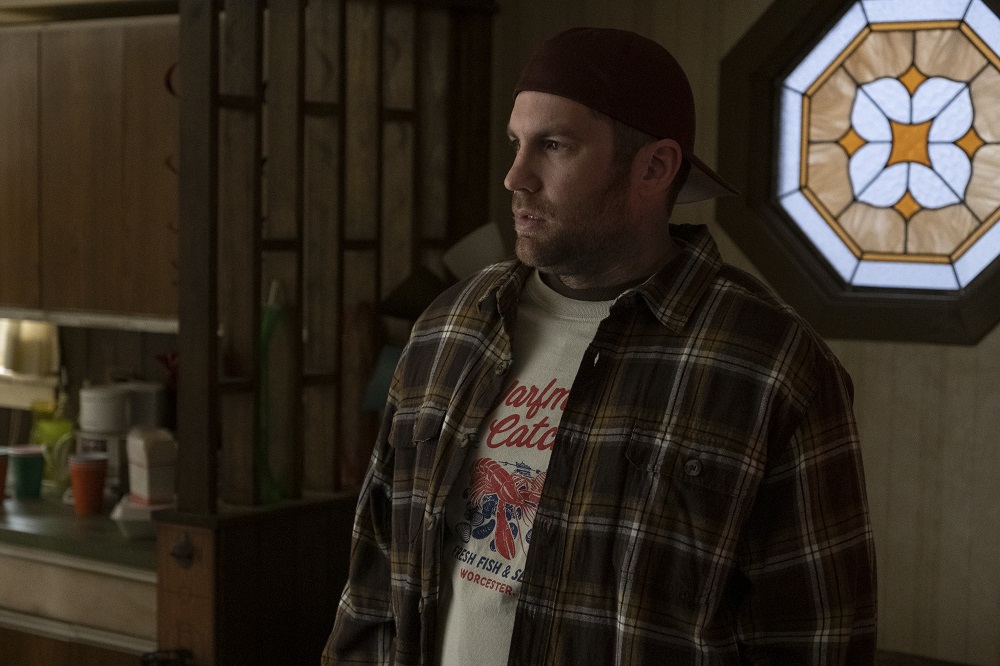 Neil stands in his apartment while wearing a white t-shirt and plaid jacket in Kevin Can F**k Himself Season 2 Episode 6, "The Machine."