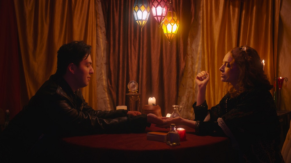 Ezra sits across the table from Somber Wales in a dimly lit room with a golden velvet curtain in EZRA.