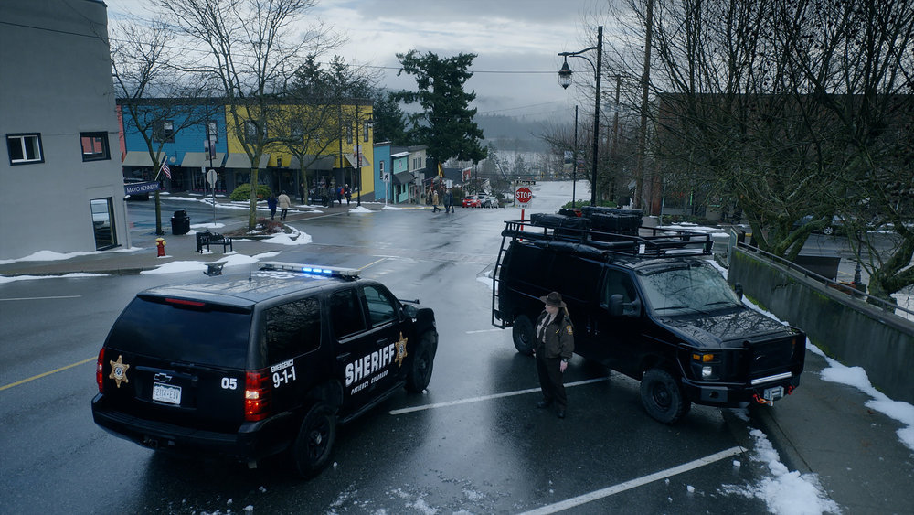 Two black police SUVs sit in an empty parking lot on an overcast day in Resident Alien Season 2 Episode 16, "I Believe in Aliens."
