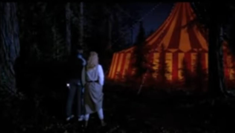 A young couple stand in front of a yellow circus tent in the middle of the woods