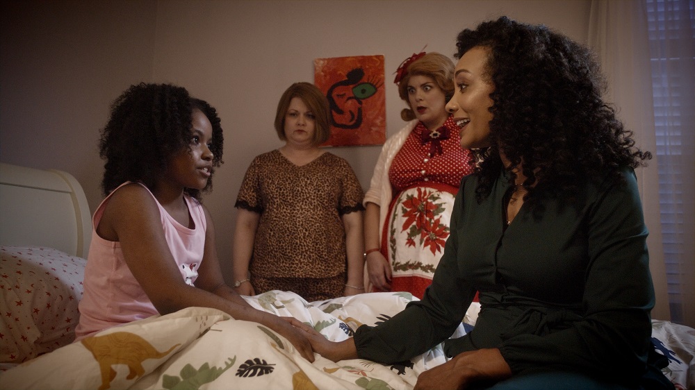 Michele Simms as Karen and Leyla Lawrence as Joy, the Ghost of Christmas Present, watch on while Robin (Ashley Jones) talks with her daughter Nia (Amina Massai) on a bed in A Christmas Karen.