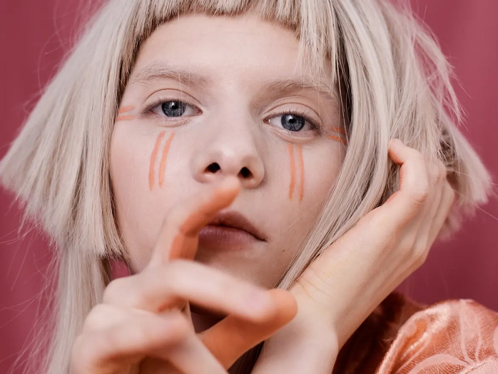 Promotional shot of Norwegian singer AURORA, including a closeup of her face on a red backdrop.