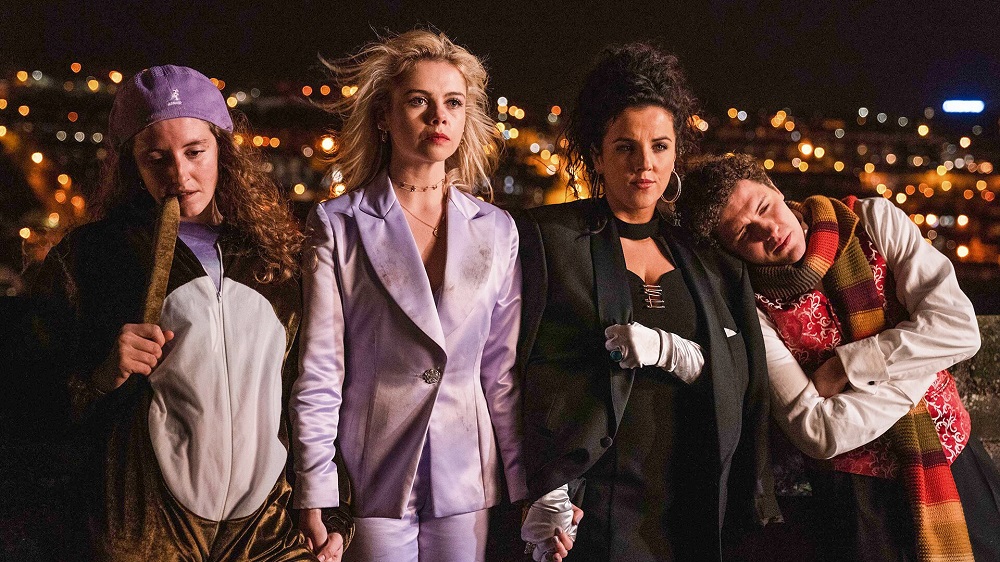 Orla, Erin, Michelle and James stand outside at night while leaning on each other in front of a stone wall in Derry Girls Season 3.