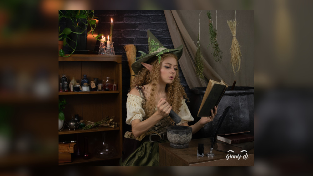 Ginny Di dressed in a wood elf witch like cosplay reading from a spell book and mixing ingreditents. 
