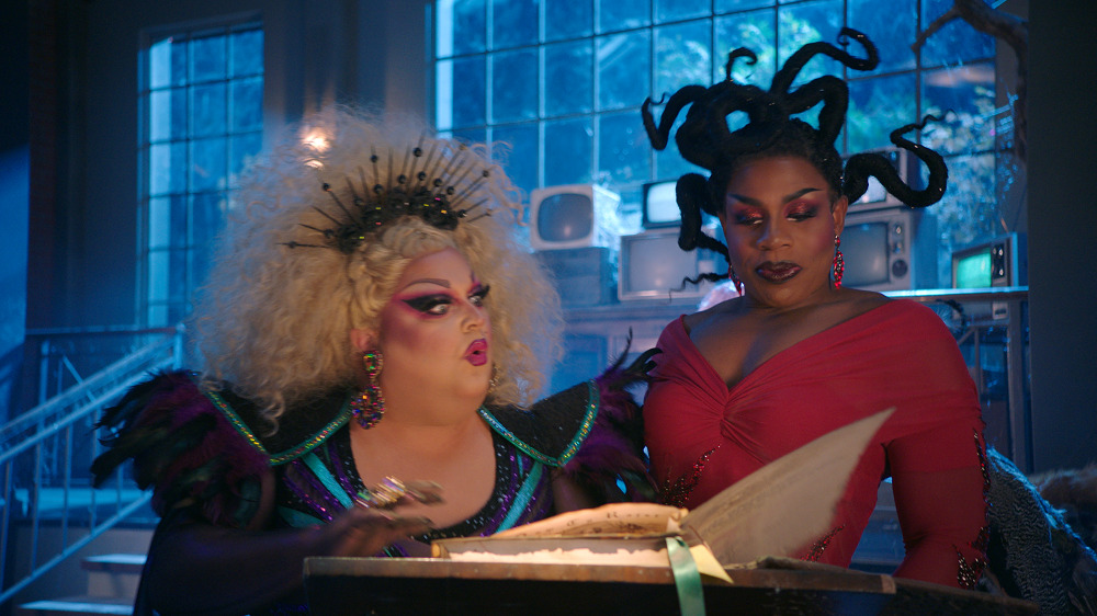 Ginger Minj and Monet X Change flip through the official Huluween book while looking pensive in Huluween Dragstravaganza.