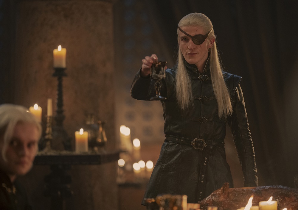 Aemond Targaryen stands by a dinner table while holding a glass of wine to toast someone in House of the Dragon Season 1 Episode 8, "The Lord of the Tides."