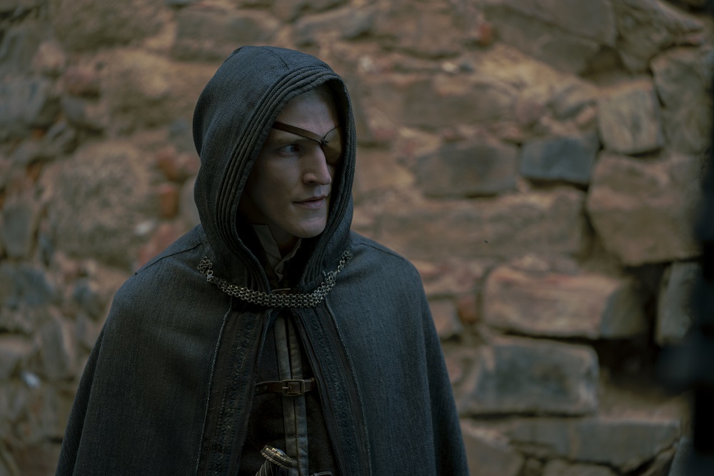 Aemond Targaryen stands in a black hooded cloak with a brick wall behind him in House of the Dragon Season 1 Episode 9, "The Green Council."