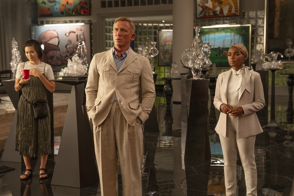 Benoit Blanc, Andi Brand, and Peg stand in a room in Miles Bron's lavish mansion while looking intently at something off-screen in Glass Onion: A Knives Out Mystery.