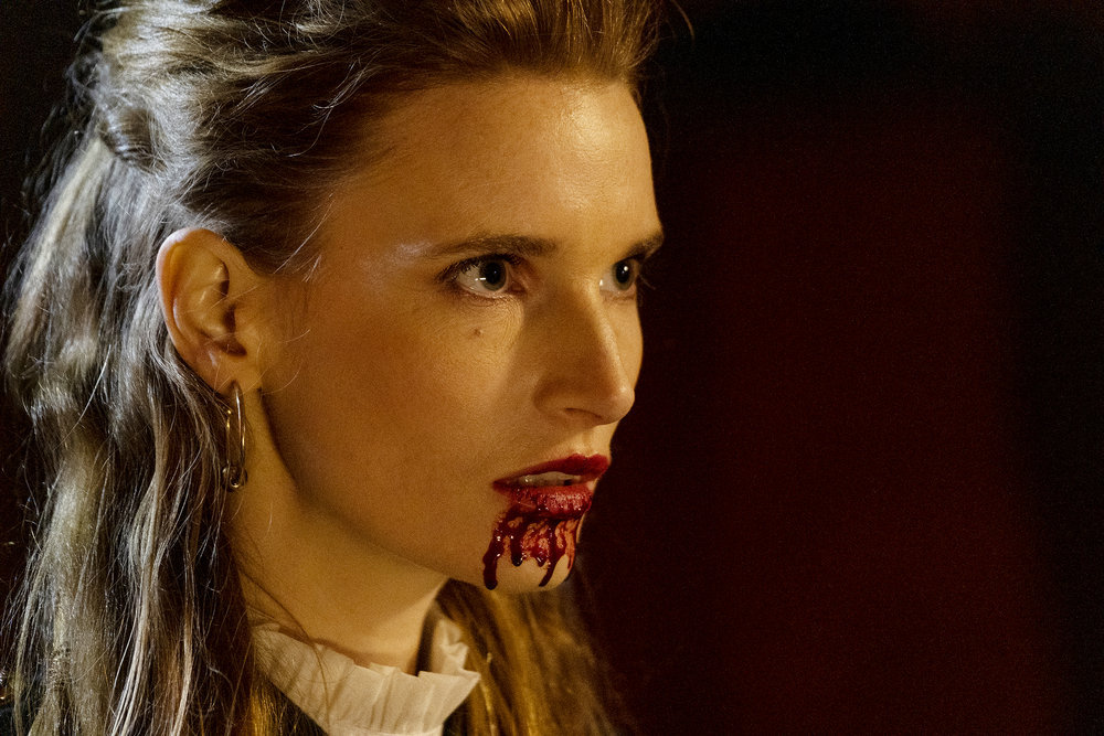 A close-up of Penelope's face (Georgia Waters) with her fangs extended and blood trickling down her chin on Reginald the Vampire.