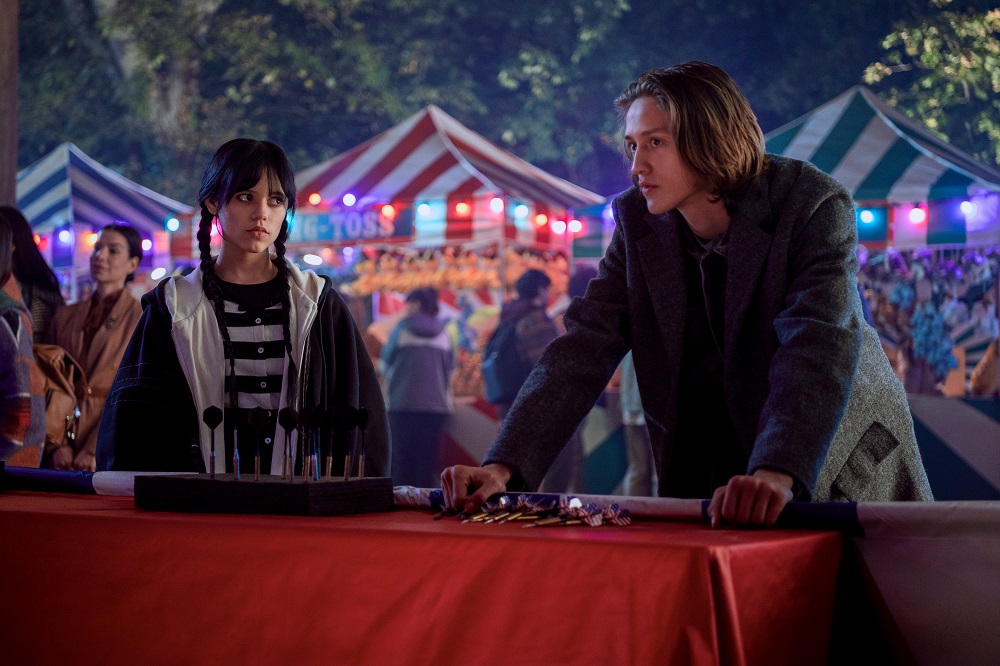 Wednesday Addams and Xavier Thorpe stand next to each other at a fair at night.
