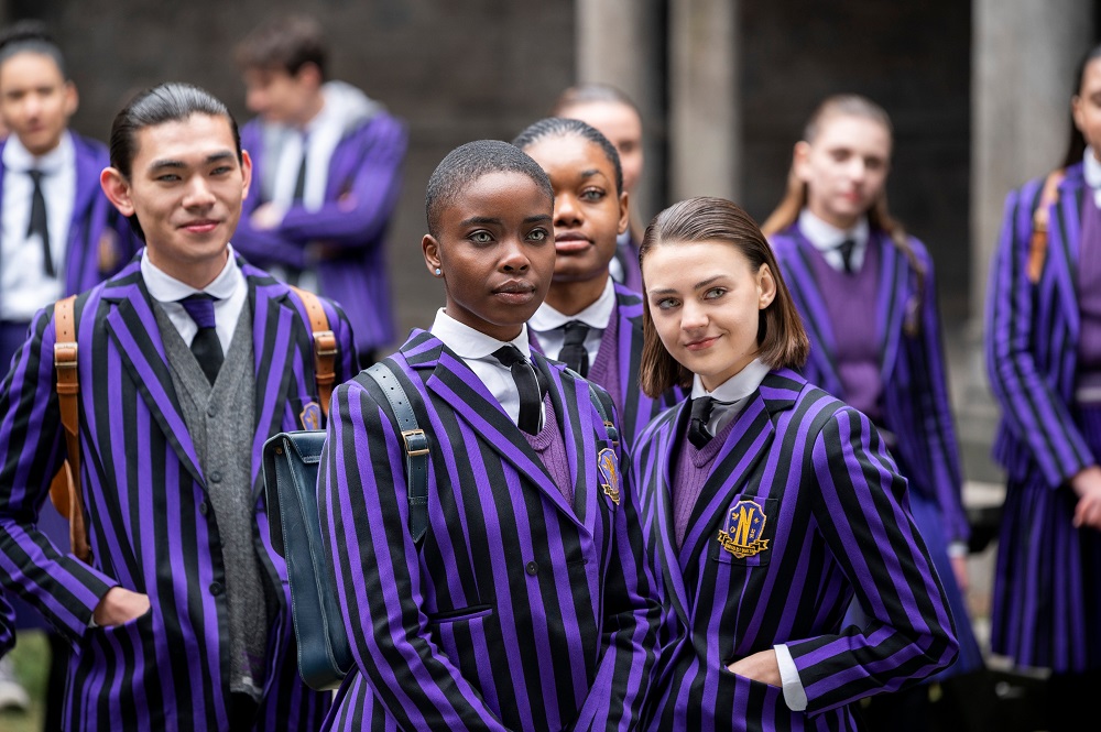Bianca stands with her group while wearing the black and purple-striped uniform of Nevermore Academy in Wednesday Season 1 Episode 3, "Friend or Woe."