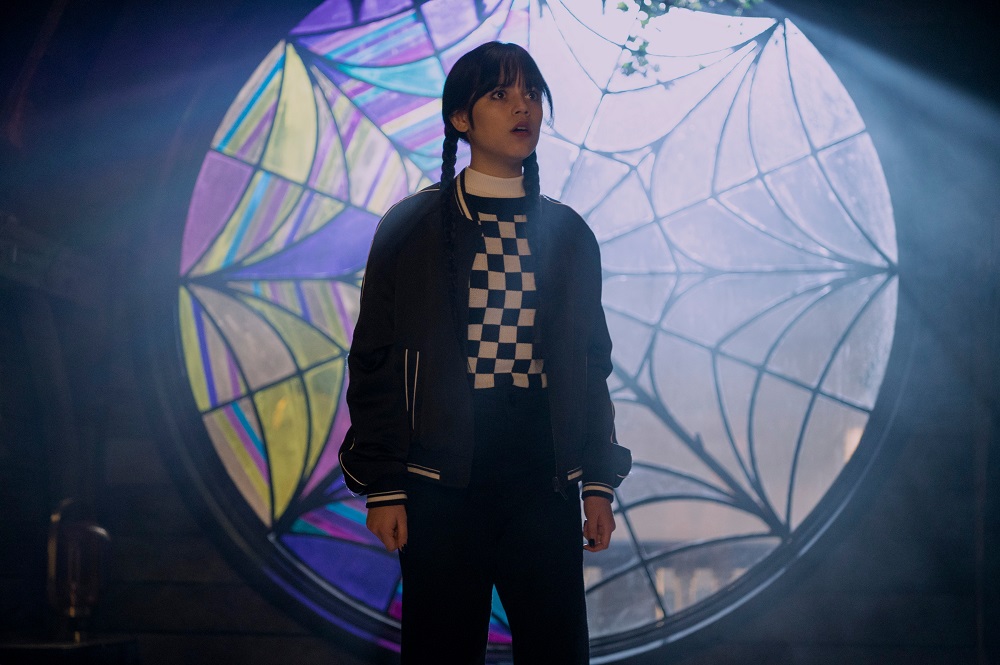 Wednesday Addams wears a black and white checkered shirt while standing in front of a stained glass window.
