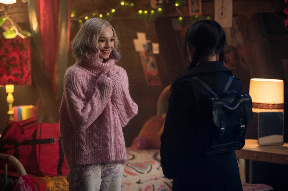 Enid Sinclair stands in front of Wednesday Addams while wearing a pink fuzzy sweater and smiling.