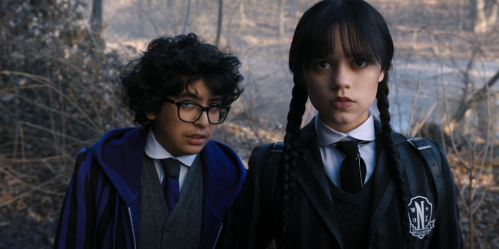 Eugene Otinger and Wednesday Addams stand in the woods in daylight while looking focused.