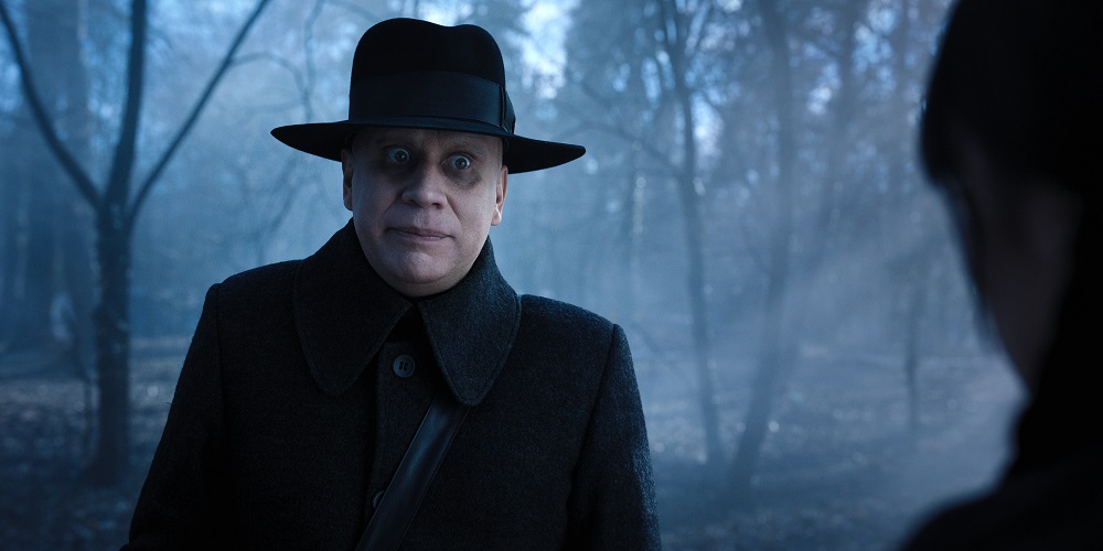 Uncle Fester wears a black hat and black coat while looking surprised in the woods in Wednesday Season 1 Episode 7, "If You Don't Woe Me by Now."