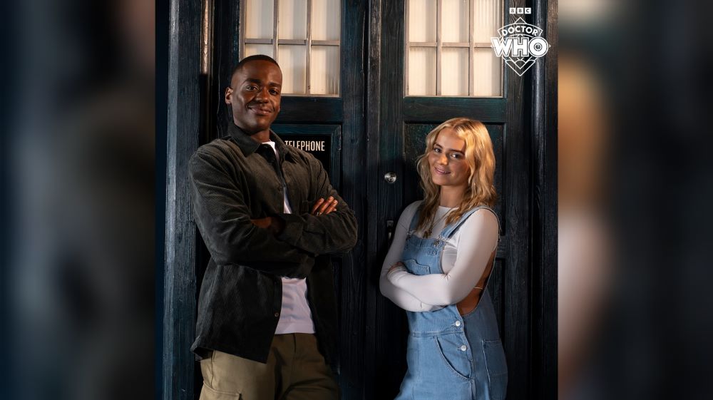 Doctor Who stars Ncuti Gatwa and Millie Gibson stand in front of the TARDIS while smiling and crossing their arms.