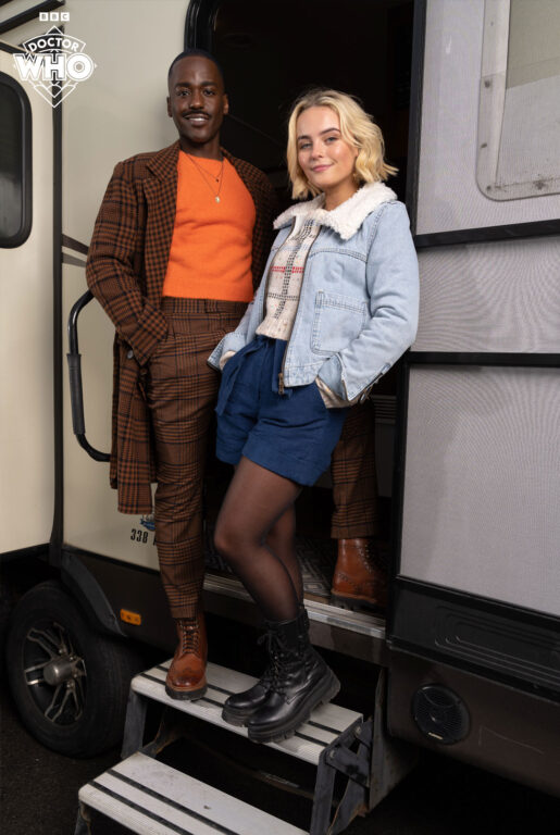 Ncuti Gatwa is dressed as the Fifteenth Doctor in a brown striped long coat and matching pants while Millie Gibson is dressed as Ruby Sunday with a light denim coat, dark blue shorts, and black tights with black combat boots.