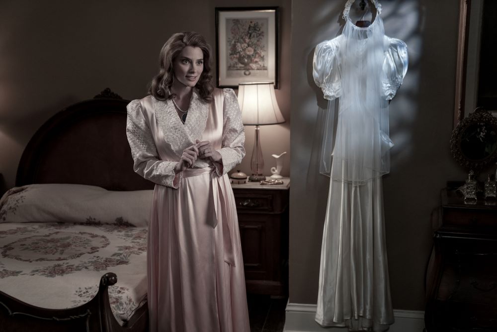 Rita stands in a bedroom while wearing a pink robe with a white wedding gown hanging on the closet door in Doom Patrol Season 4 Episode 3, "Nostalgia Patrol."