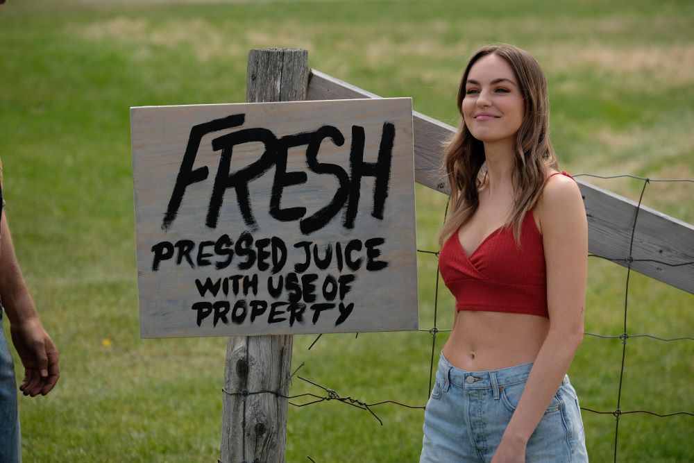 Katy, played by Michelle Mylett, stands outside on a sunny day next to a sign that says, "Fresh pressed juice with use of property" in Letterkenny Season 11 Episode 5, "Influenzas."