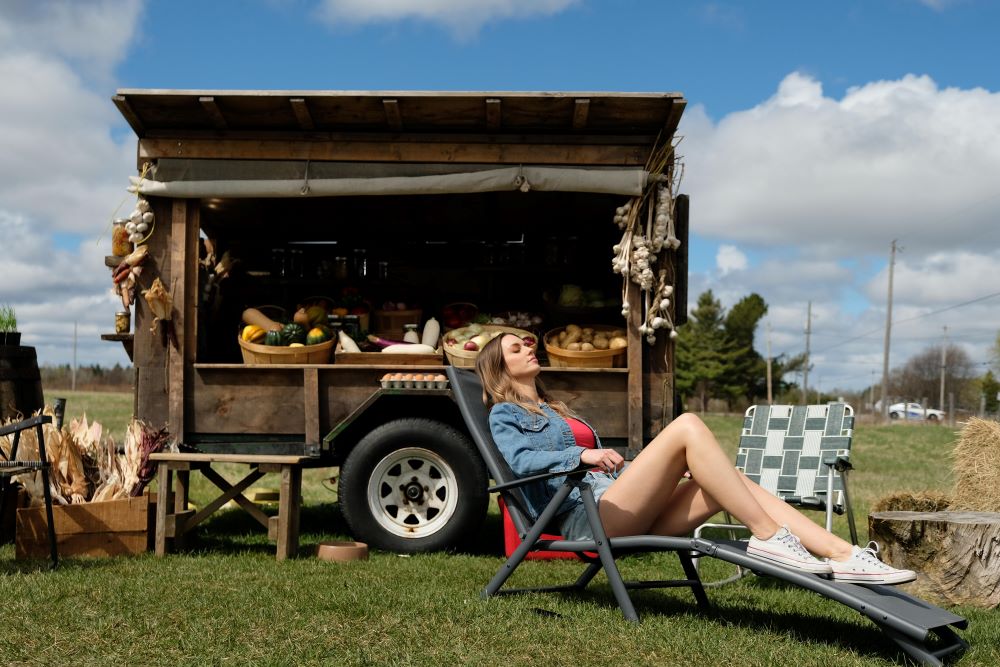 Katy lounges in a reclining chair near the fruit stand outside in Letterkenny Season 11 Episode 2, "Okoya."