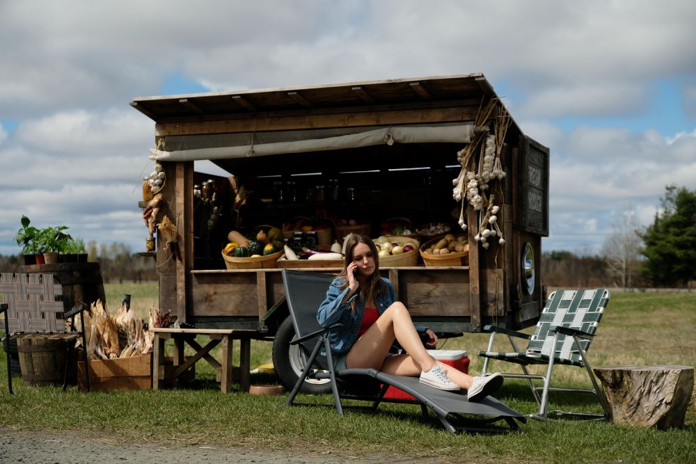 Katy sits in a chair outside near the fruit stand while talking on the phone in Letterkenny Season 11 Episode 2, "Okoya."