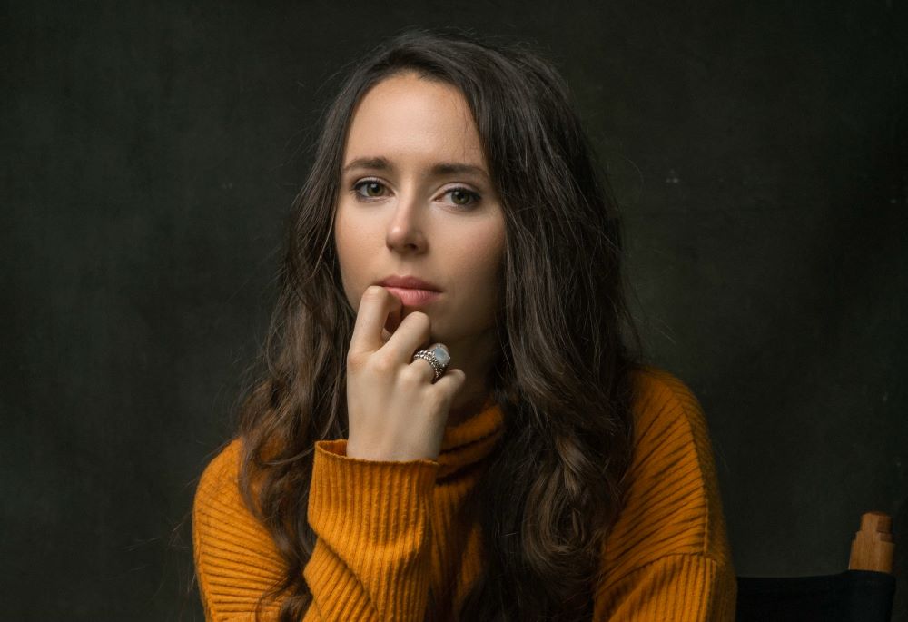 Urban Legend director Cat Hostick poses in front of a dark gray background with her hand on her chin while wearing a yellow sweater.