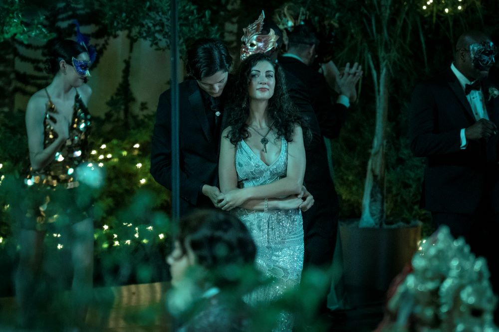 Young Deirdre wears a silver sparkling gown and stands in an outdoor party while Patrick embraces her from behind in Mayfair Witches Season 1 Episode 1, "The Witching Hour."