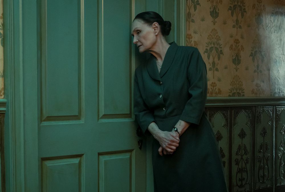 Carlotta wears a dark green dress while standing outside a bedroom door with her hands folded in front of her in Mayfair Witches Season 1 Episode 1, "The Witching Hour."